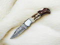 Handmade Damascus folding knife with stag horn handle and rosewood accents, showcasing intricate blade craftsmanship.