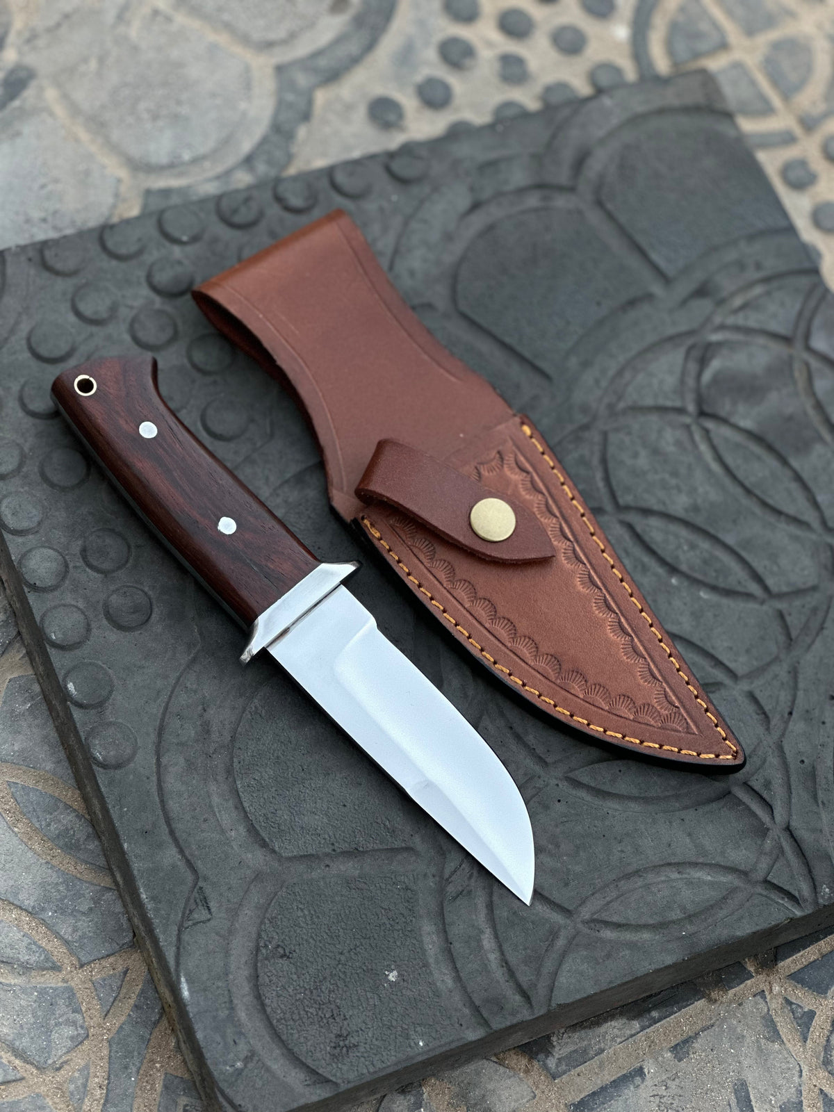 Handmade 1095 steel hunting knife with a leather sheath and wooden handle, displayed on a rustic background.