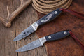 Cowboy knives from cskforged Rancher Buckaroo Collection with 3-inch Damascus blade and belt sheath, displayed on a wooden surface.