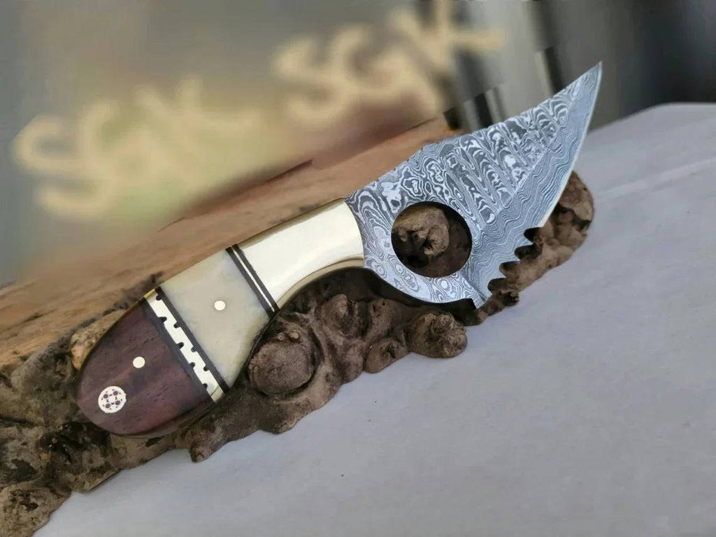 Damascus steel EDC skinning knife with finger hole, bone-rose wood handle, brass bolster, and serrated edge.