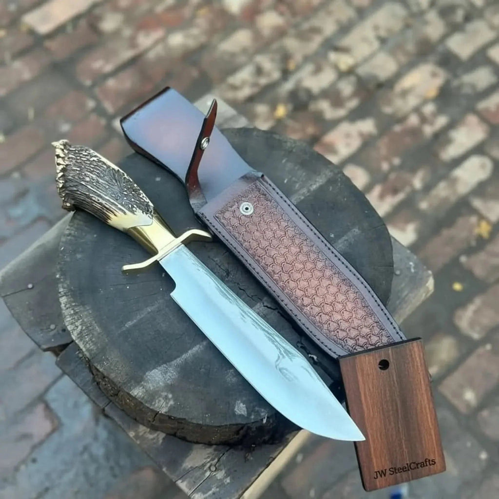Handmade JW SteelCrafts carbon steel hunting knife with stag horn handle and leather sheath, displayed on a wooden surface.