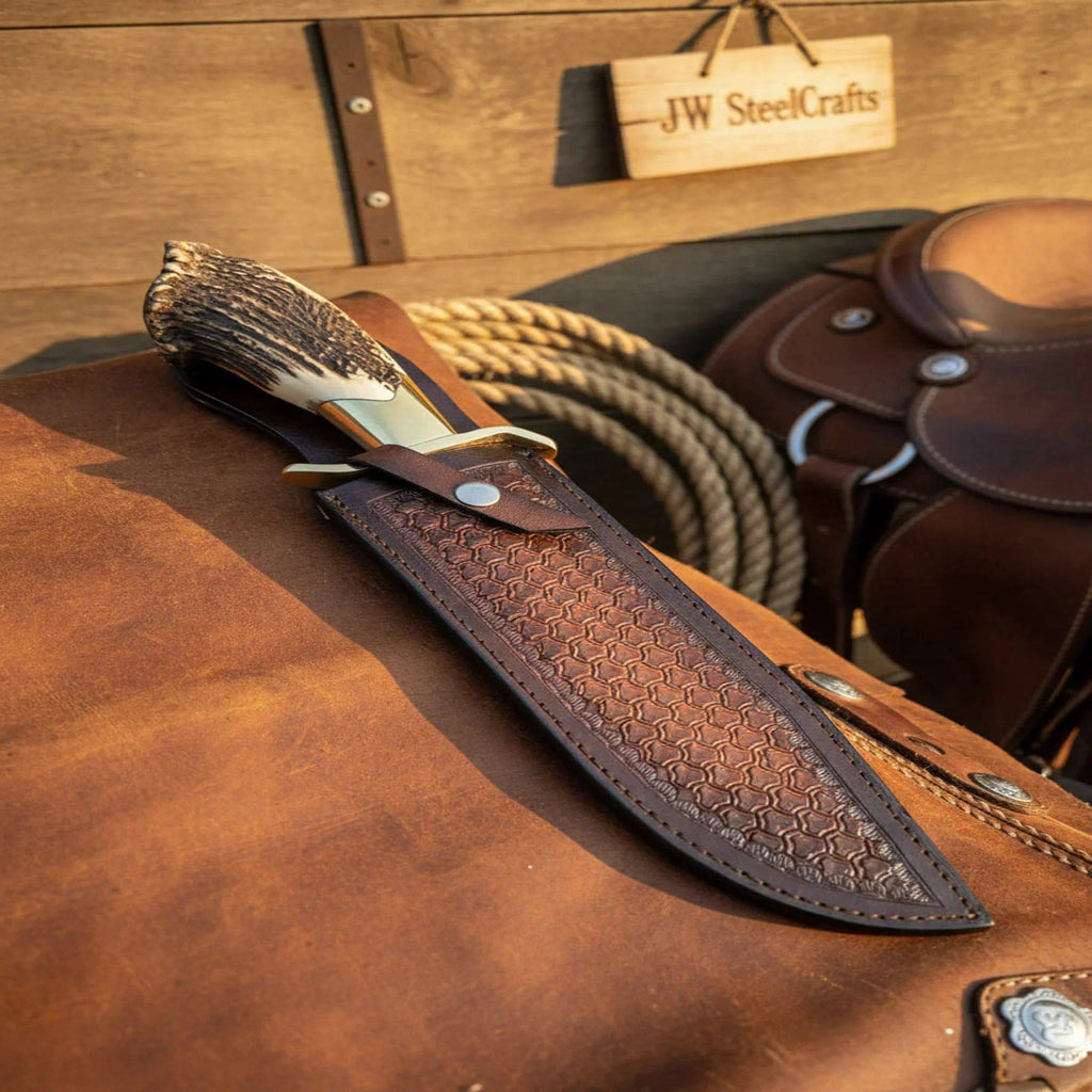 Handmade JW SteelCrafts carbon steel hunting knife with stag horn handle and leather sheath, displayed on a wooden surface.