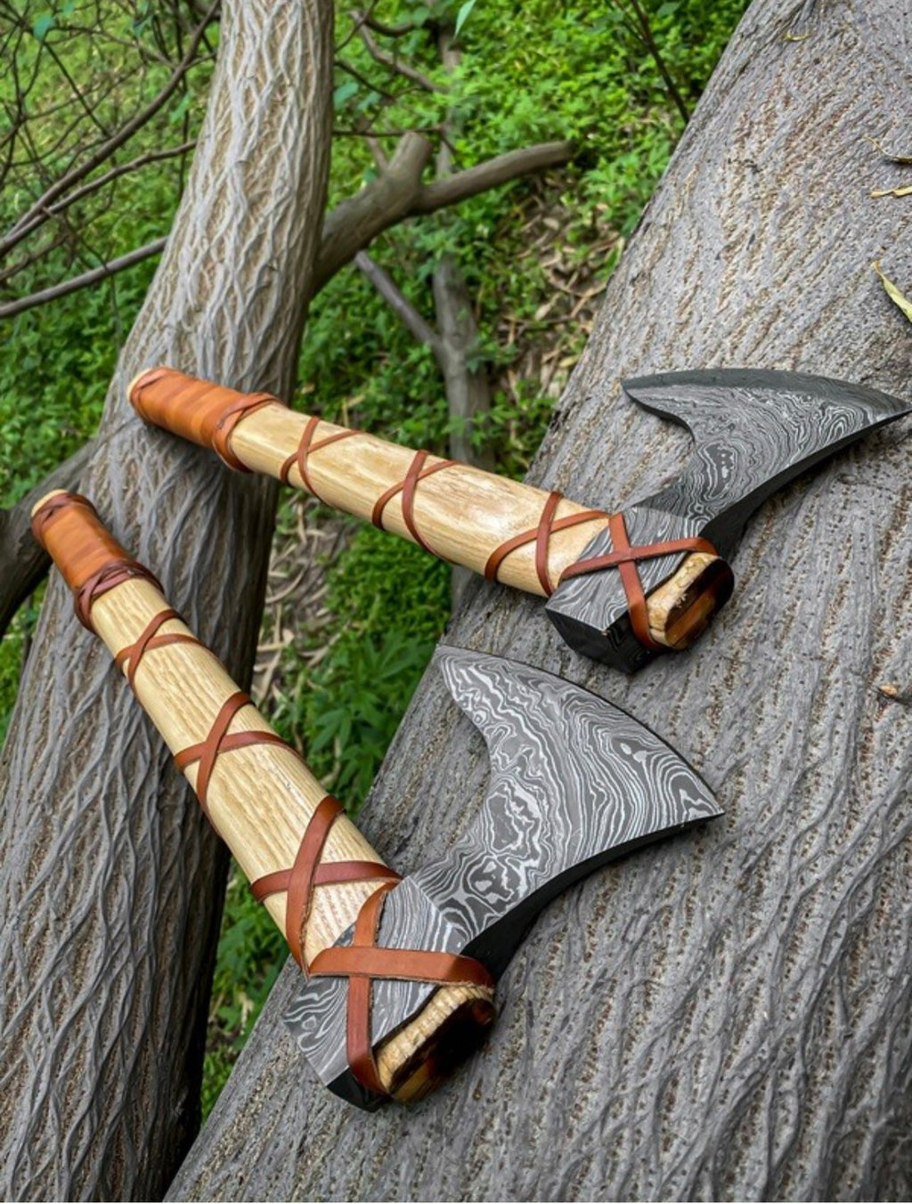 Pair of Damascus Viking axes with ashwood handles, leather wraps, and sheaths, featuring detailed metal blades and traditional craftsmanship.