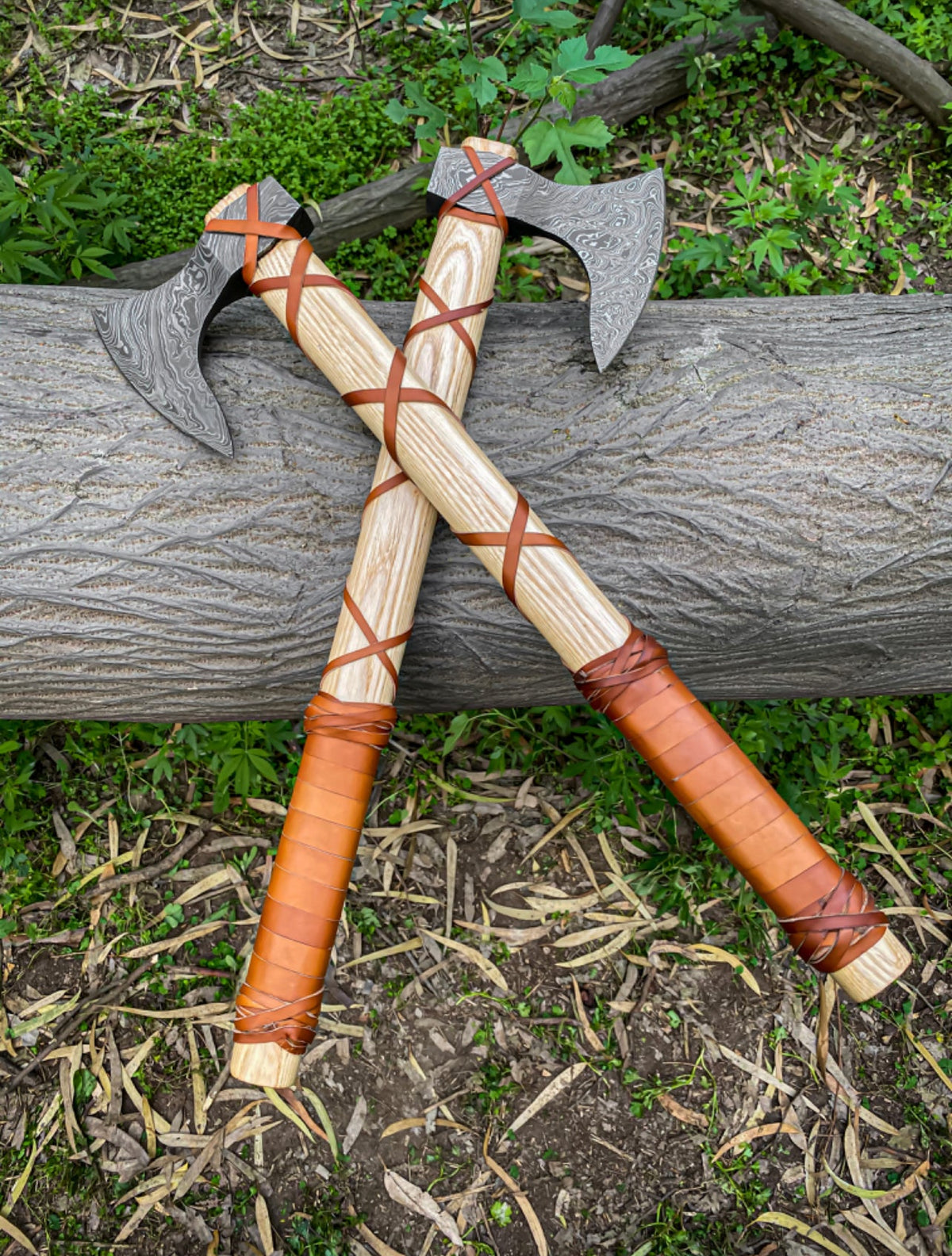 Pair of Damascus Viking axes with ashwood handles, leather wraps, and sheaths, featuring intricate blade patterns and sturdy craftsmanship.
