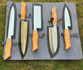 Set of Knives Gear Garage D2 steel forged kitchen knives with natural wood handles, mosaic pins, and leather sheaths displayed on a countertop.