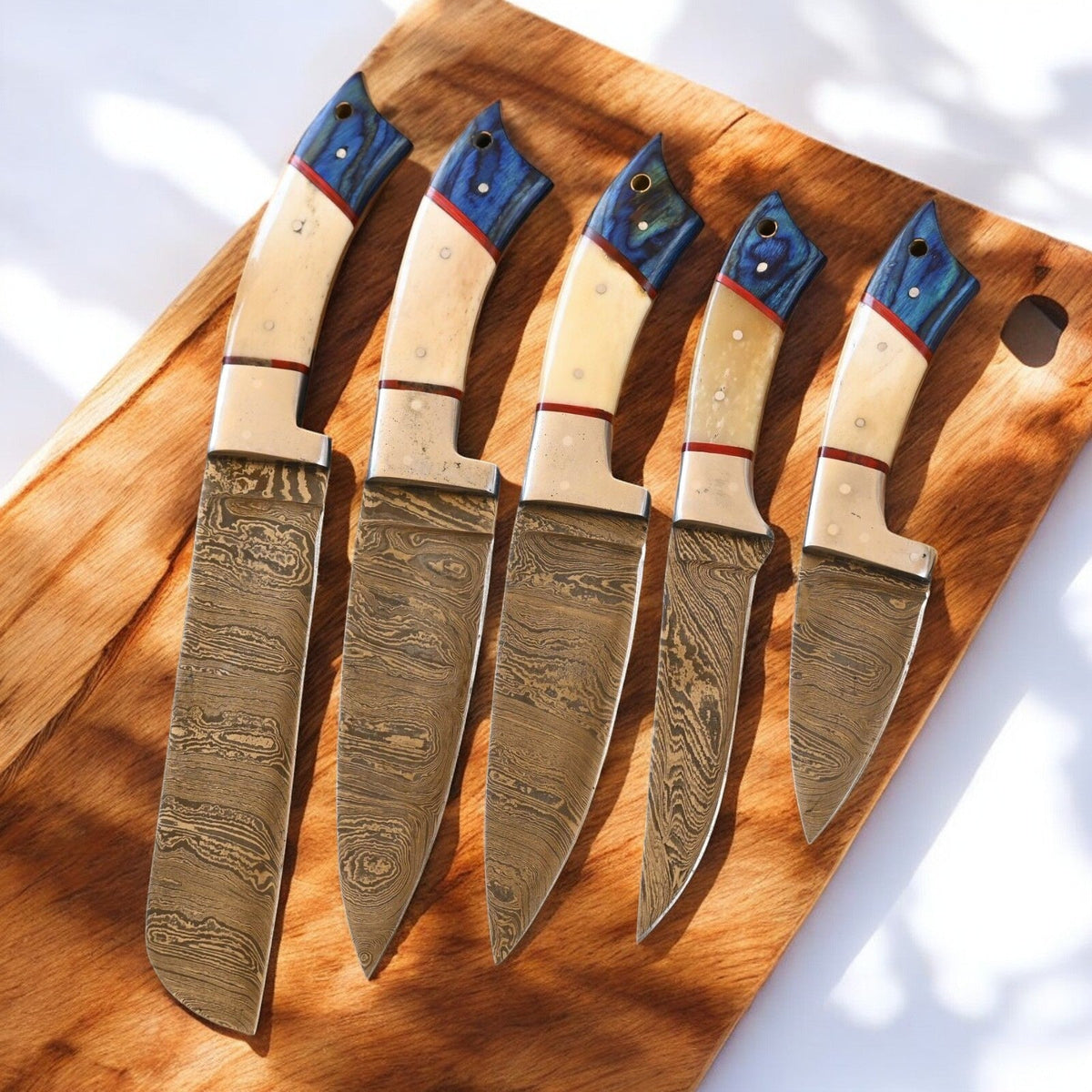 Handmade Damascus kitchen knives set with exotic wood handles and bone accents, displayed on a wooden surface.